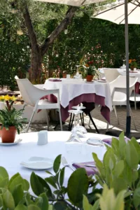 Terraza exterior de Le Gourmand en Jávea con mesas vestidas de blanco, servilletas malva y sillas modernas bajo olivos y vegetación mediterránea